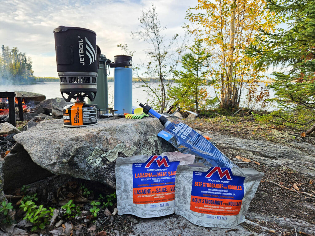 A Jetboil and Mountin House meals sit on a rock by a lake